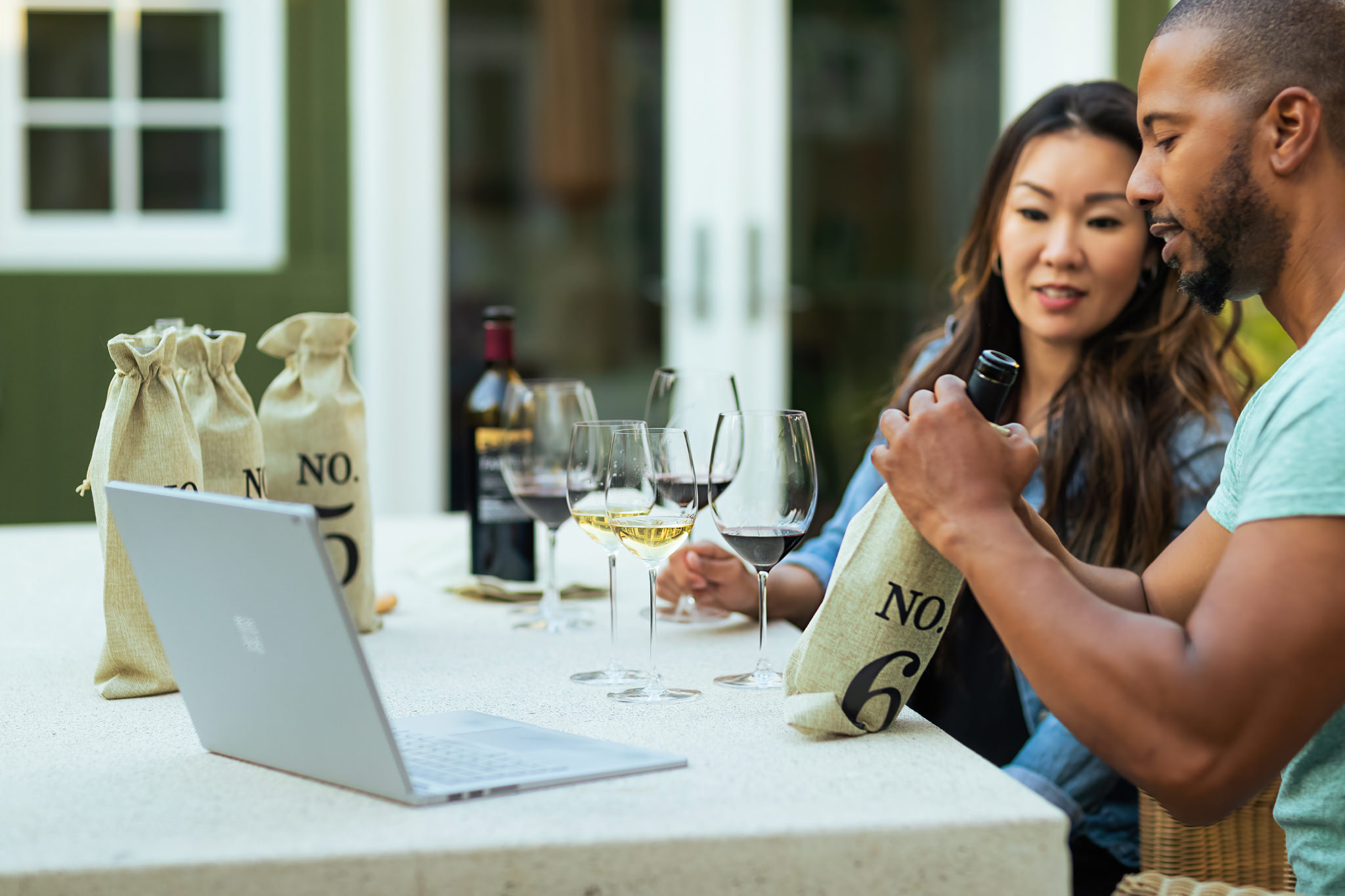 couple outside tasting wine from numbered bottles in bags and watching laptop
