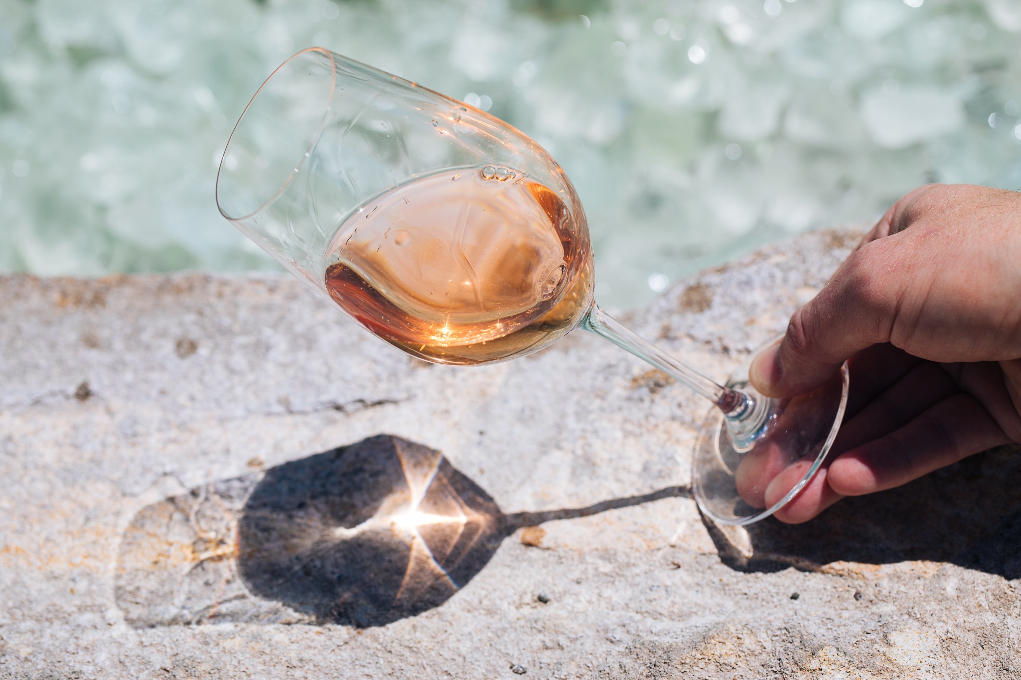 male hand holding a glass of rosé wine at an angle in front of a water feature